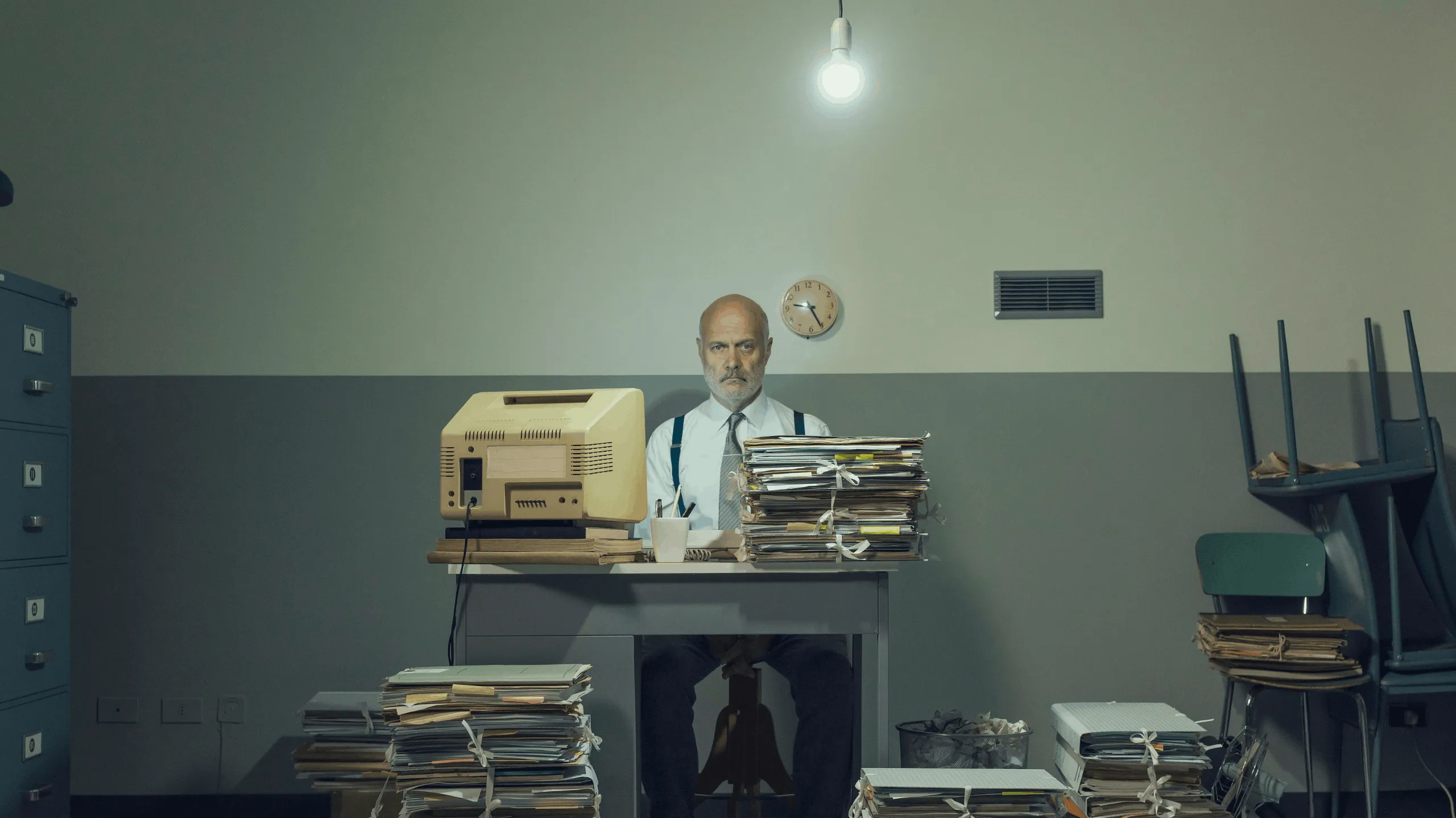Man in suit with outdated IT solutions including old computer and files sits at desk in 70s style office